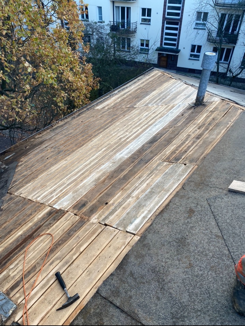 Widok na drewnianą konstrukcję dachu płaskiego w trakcie prac, widoczne deski, papa i narzędzie - młotek dekarski, w tle budynek mieszkalny i drzewa.