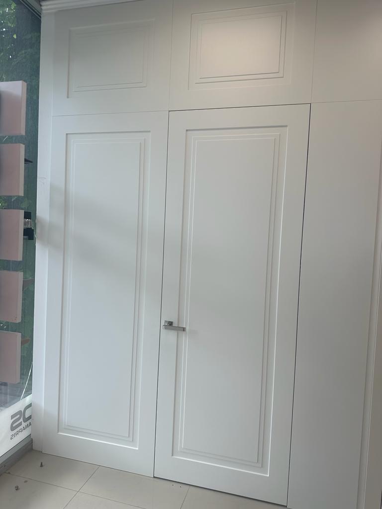 Białe, dwuskrzydłowe drzwi wewnętrzne z subtelnymi frezowaniami w ramkach, zamknięte, z chromowaną klamką, stanowiące część zabudowy meblowej sięgającej sufitu, widoczne z bliska, z elementami...