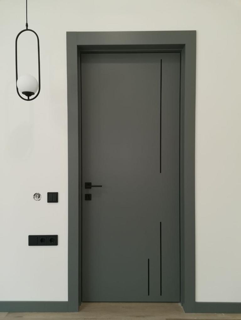 Szare drzwi z czarnymi, pionowymi intarsjami, czarna klamka i ościeżnica w odcieniu szarości, na ścianie czarne gniazdka i włączniki, minimalistyczna lampa wisząca z białą kulą.