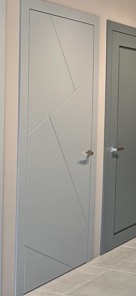 Szare skrzydło drzwiowe z geometrycznymi frezowaniami, widoczny fragment drugiego skrzydła i podłogi z płytek.