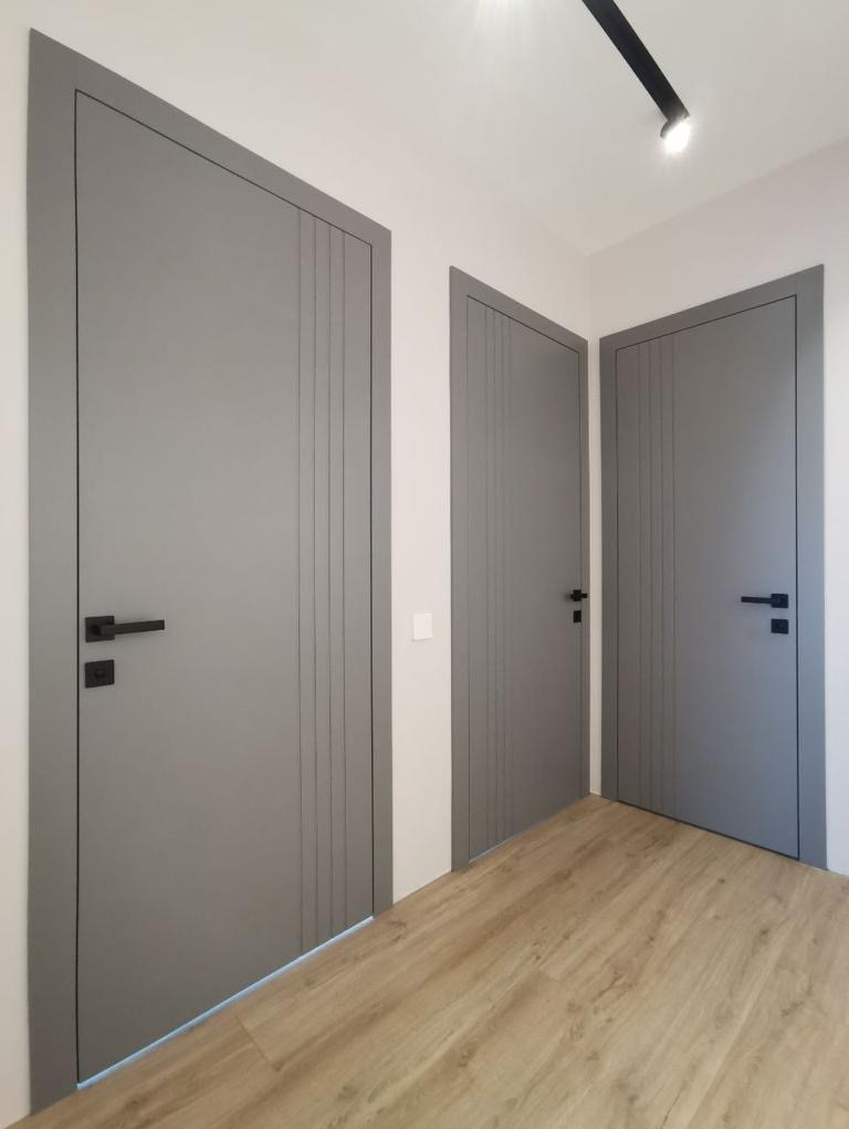 Trzy szare skrzydła drzwiowe z czarnymi klamkami, pionowymi frezowaniami i cienką obwódką w nowoczesnym wnętrzu z panelami podłogowymi.