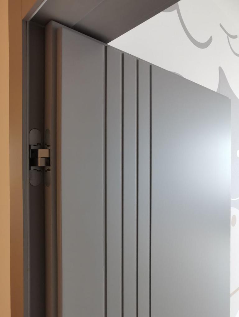 Szare, pionowo żłobkowane skrzydło drzwiowe z widocznym zawiasem i fragmentem dekoracyjnej tapety w tle.