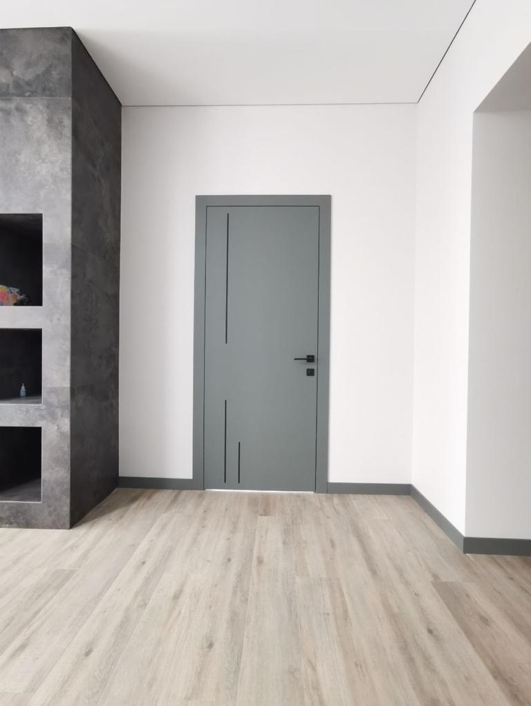 Szare drzwi wewnętrzne z minimalistycznymi, pionowymi frezowaniami, zamontowane w białej ścianie z szarą listwą przypodłogową, widok z perspektywy podłogi z paneli w odcieniach beżu.