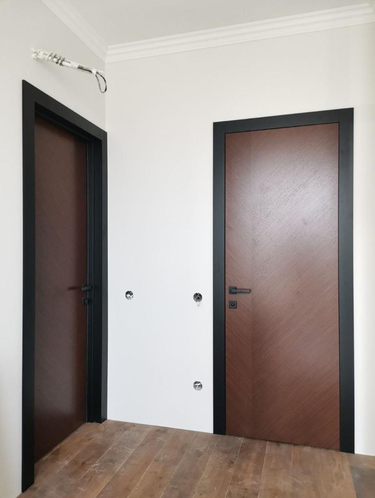 Dwa brązowe skrzydła drzwiowe z czarnymi ościeżnicami zamontowane w białej ścianie z widocznymi puszkami elektrycznymi, drewniana podłoga.