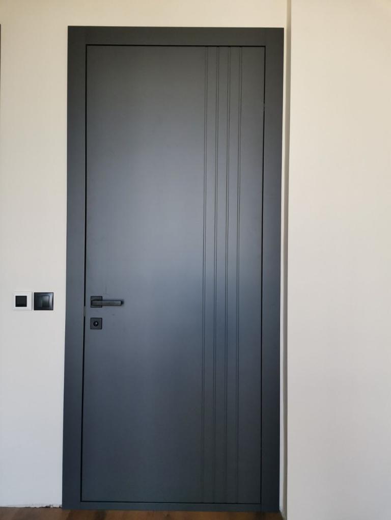 Grafitowe skrzydło z pionowymi frezami i czarną klamką na tle jasnej ściany z włącznikami.