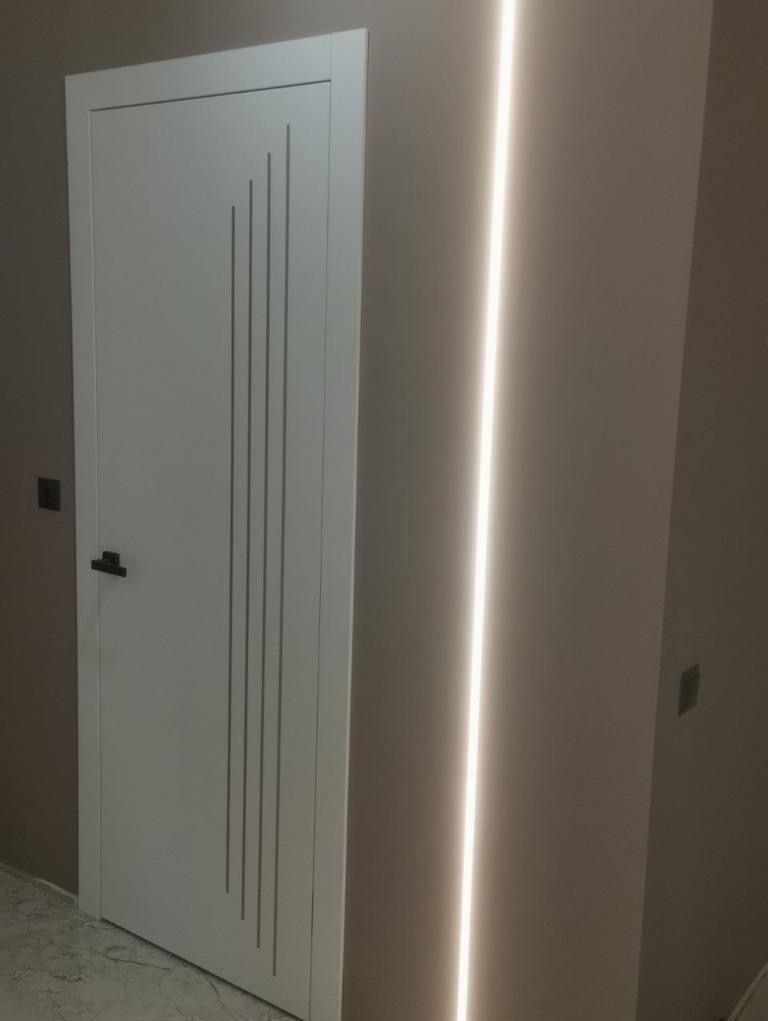 Białe drzwi wewnętrzne z pionowymi, asymetrycznymi żłobieniami, zamontowane w ścianie z pionowym oświetleniem LED, widoczne z bliska.