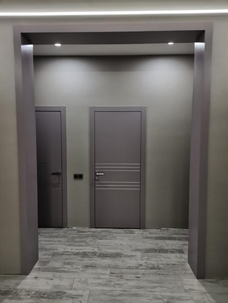 Dwa skrzydła drzwiowe w szarych odcieniach, widoczne przez szarą framugę, na podłodze imitacja drewna, oświetlenie sufitowe LED.