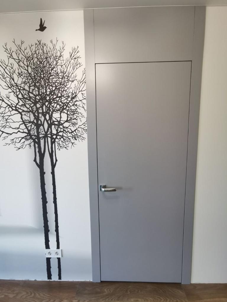 Szare, bezprzylgowe drzwi w minimalistycznym wnętrzu z dekoracyjną naklejką drzewa i ptaka na ścianie.