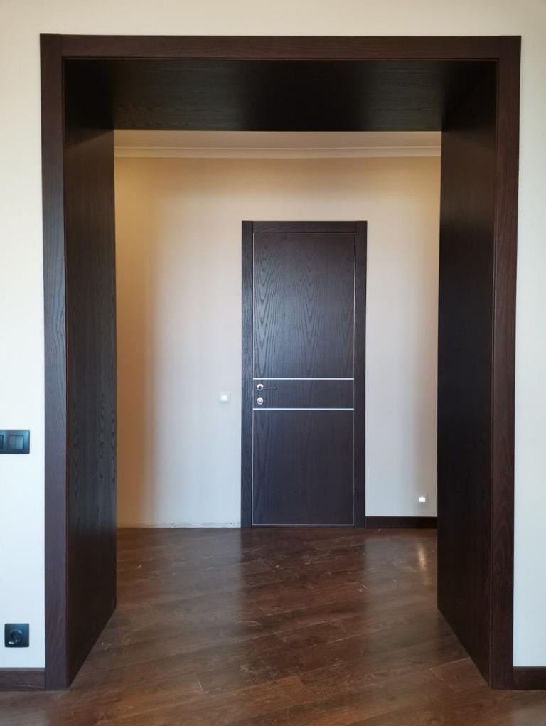 Ciemnobrązowa futryna z widokiem na zamknięte, gładkie drzwi w kolorze wenge z dwoma poziomymi, srebrnymi paskami, na tle jasnej ściany i parkietu.