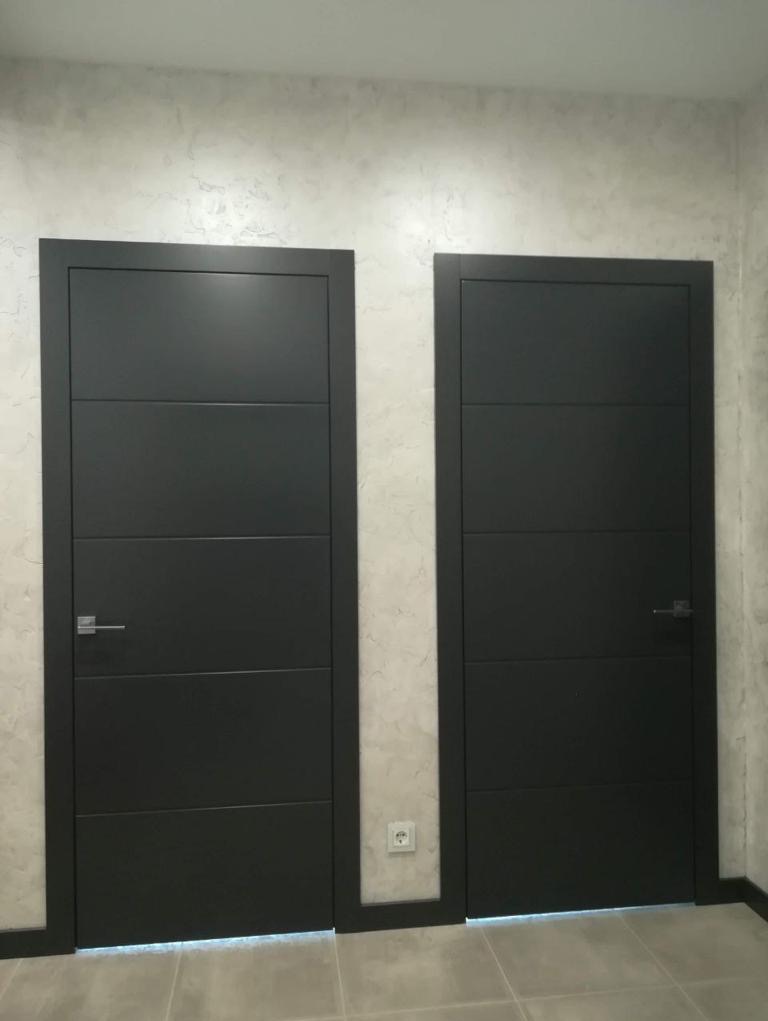 Dwa ciemnoszare skrzydła drzwiowe z poziomymi panelami w nowym mieszkaniu, widoczne gniazdko elektryczne na ścianie, szare płytki na podłodze.