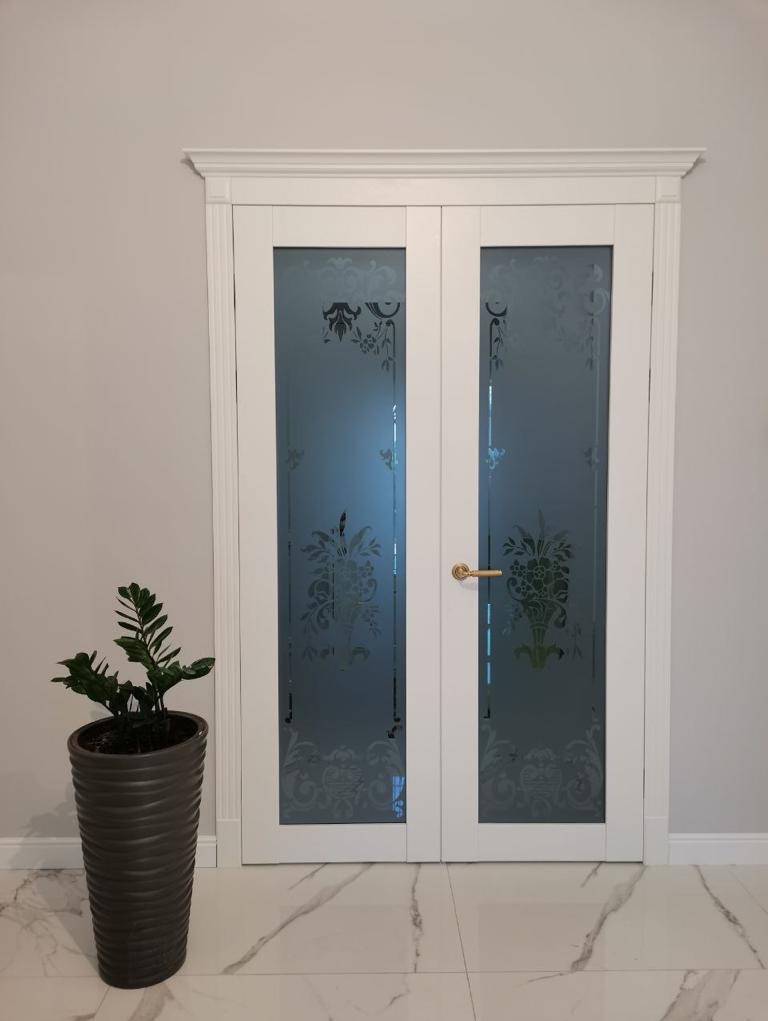 Białe dwuskrzydłowe drzwi z matowego szkła z ornamentami, złota klamka, na podłodze marmurkowy wzór, obok donica z rośliną.