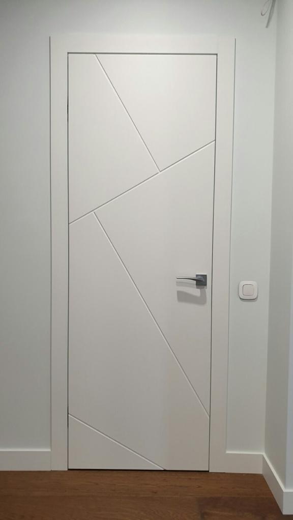 Białe drzwi z geometrycznym wzorem, srebrna klamka, widoczny włącznik światła na ścianie, parkiet na podłodze.