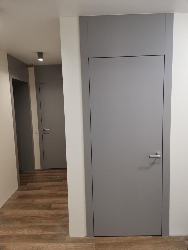 Szara, minimalistyczna ościeżnica z ukrytymi zawiasami, zlicowana ze ścianą, widoczna w korytarzu z dwiema podobnymi konstrukcjami, drewniana podłoga.