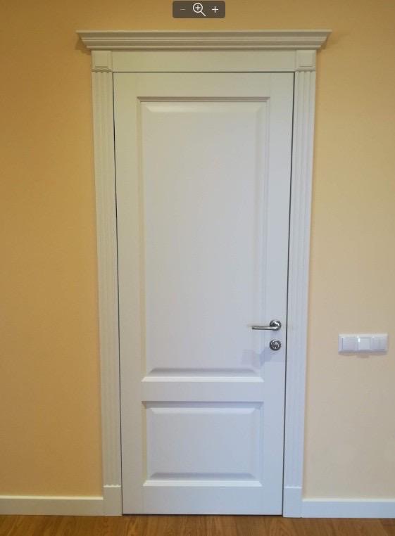 Białe, dwuczęściowe drzwi z ozdobnymi listwami i srebrną klamką, zamontowane w ścianie o jasnym, brzoskwiniowym odcieniu, widoczny włącznik światła.