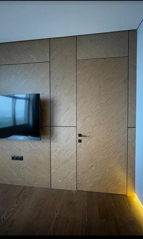 Ukryte drzwi zlicowane ze ścianą z paneli drewnopodobnych, minimalistyczna czarna klamka, telewizor na ścianie, oświetlenie podłogowe LED.