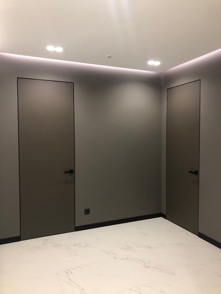 Dwie minimalistyczne, ukryte drzwi z czarnymi ramami i klamkami, w szarej ścianie, oświetlenie LED podsufitowe, biała marmurowa podłoga, widoczny mały włącznik na ścianie.