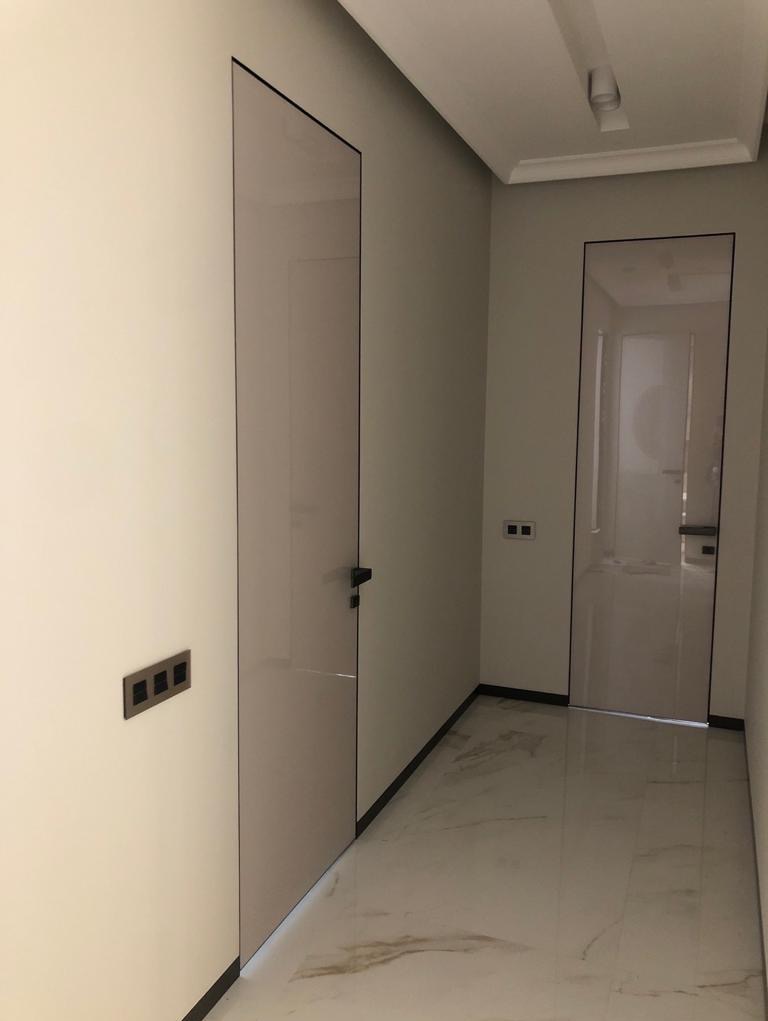 Nowoczesny korytarz z dwiema ukrytymi, bezprzylgowymi drzwiami zlicowanymi ze ścianą, wykończonymi w odcieniach bieli i szarości, z widocznymi czarnymi listwami przypodłogowymi i marmurową podłogą...