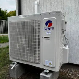 Zamontowana jednostka zewnętrzna klimatyzacji GREE na betonowych bloczkach przy ścianie budynku o chropowatej fakturze. Widoczne elementy instalacji i trawa.
