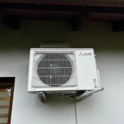 Biała jednostka zewnętrzna klimatyzacji Mitsubishi Electric zamontowana na elewacji budynku z widocznymi elementami instalacji i rynną w tle.