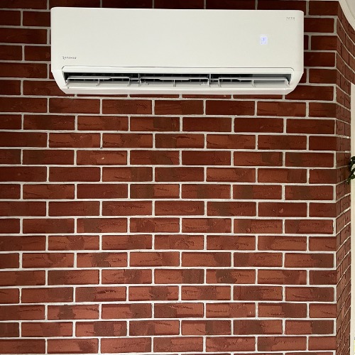 Biała klimatyzacja na ceglanej ścianie z widocznym wyświetlaczem temperatury, zamontowana w rogu pomieszczenia. Widok z dołu.