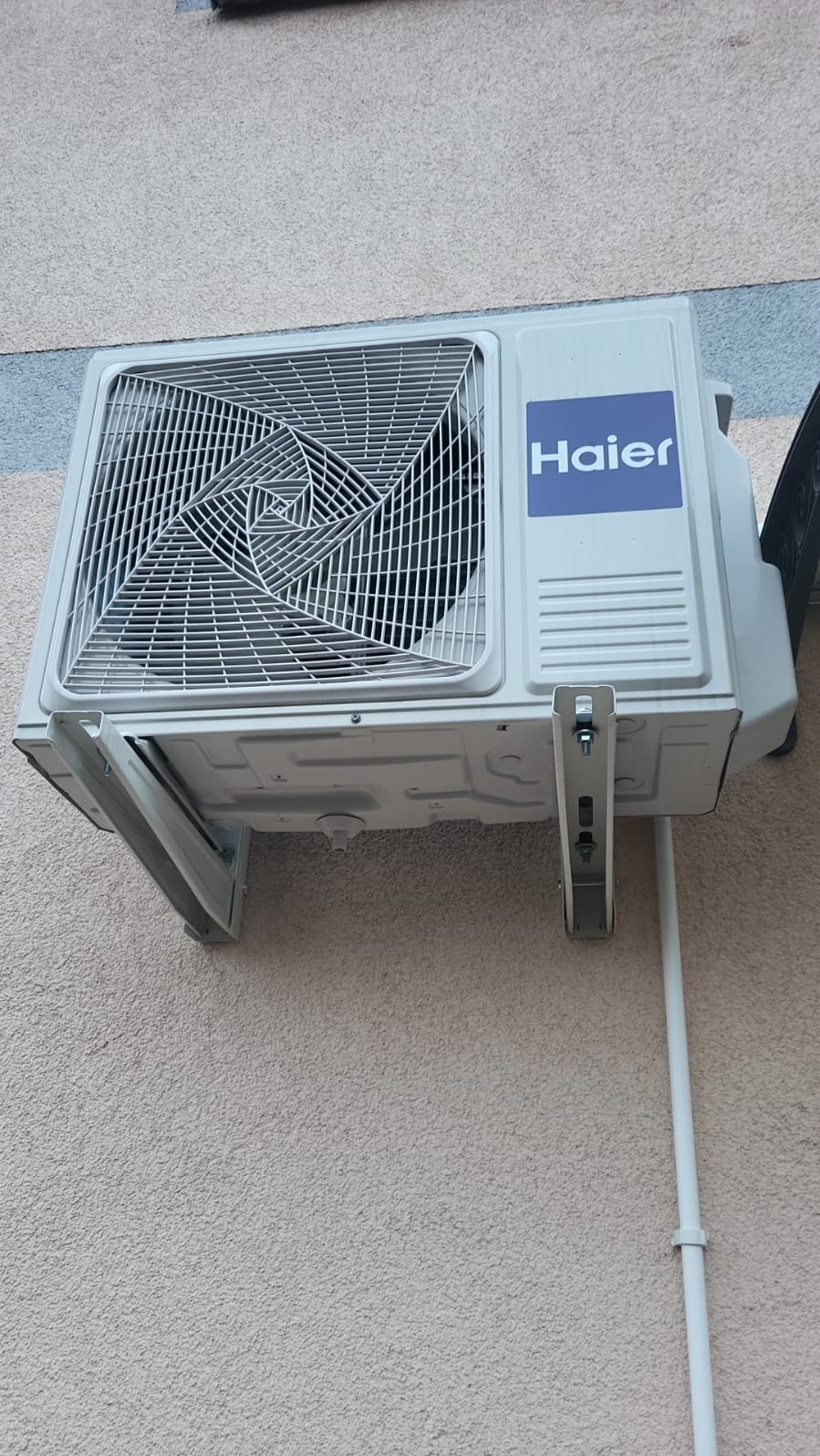 Zamontowana jednostka zewnętrzna klimatyzacji Haier na beżowej elewacji budynku, widoczne mocowania i rura odprowadzająca skropliny. Ujęcie z dołu.