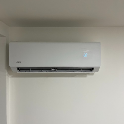 Biały klimatyzator AUX zamontowany na jasnej ścianie. Na wyświetlaczu widoczna temperatura 22 stopnie. Minimalistyczny design, widok z dołu.