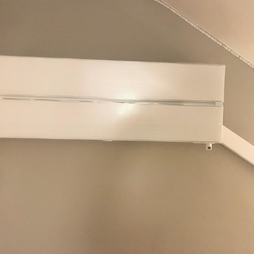 Biała jednostka wewnętrzna klimatyzacji zamontowana na ścianie. Widoczny fragment sufitu z listwą. Minimalistyczny design urządzenia.