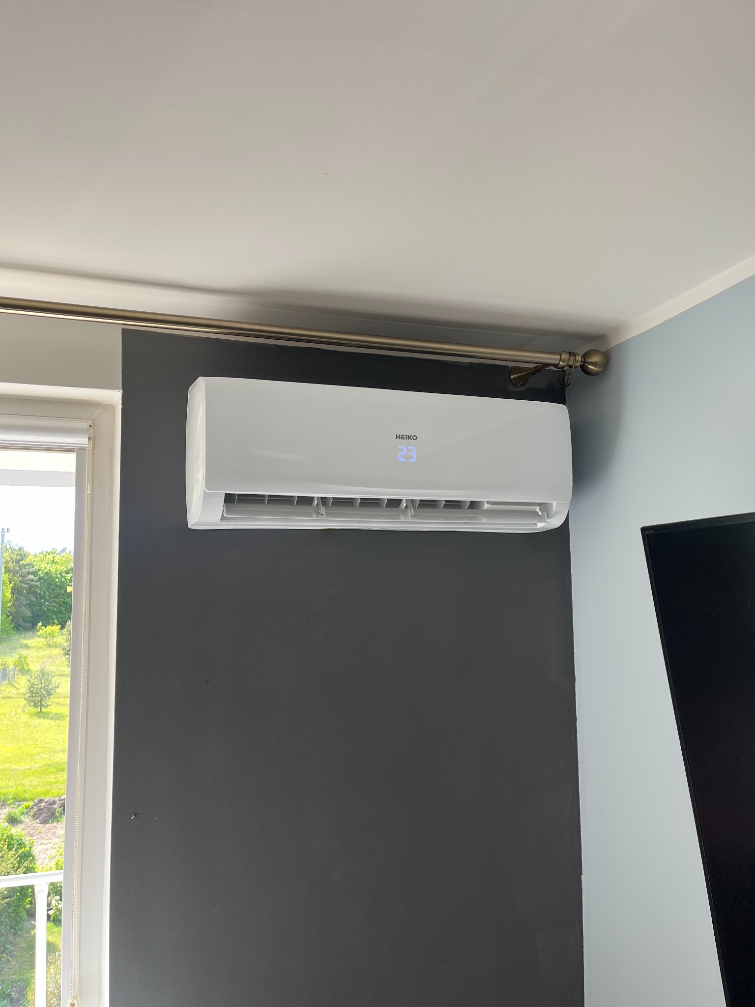 Biała klimatyzacja Heiko na szarej ścianie w pokoju z oknem i widokiem na zieleń. Na wyświetlaczu widoczna temperatura 23 stopnie.