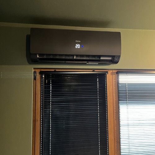 Ciemnoszara klimatyzacja Haier na ścianie w pokoju, pod oknem z żaluzjami. Na wyświetlaczu urządzenia widoczna temperatura 20 stopni.