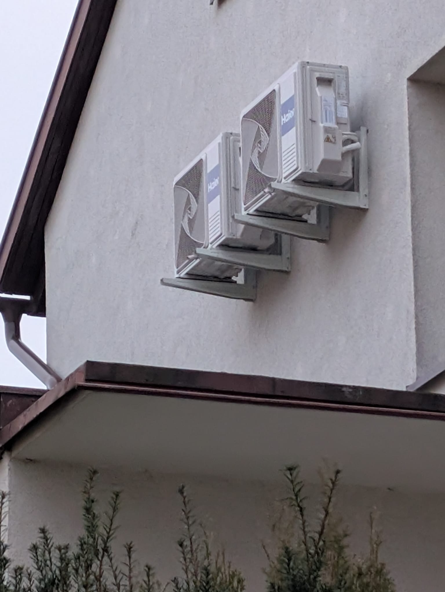 Trzy klimatyzatory Haier zamontowane kaskadowo na jasnej ścianie budynku. Widoczne elementy montażowe i fragment dachu z rynną. Tło stanowi zieleń krzewów.