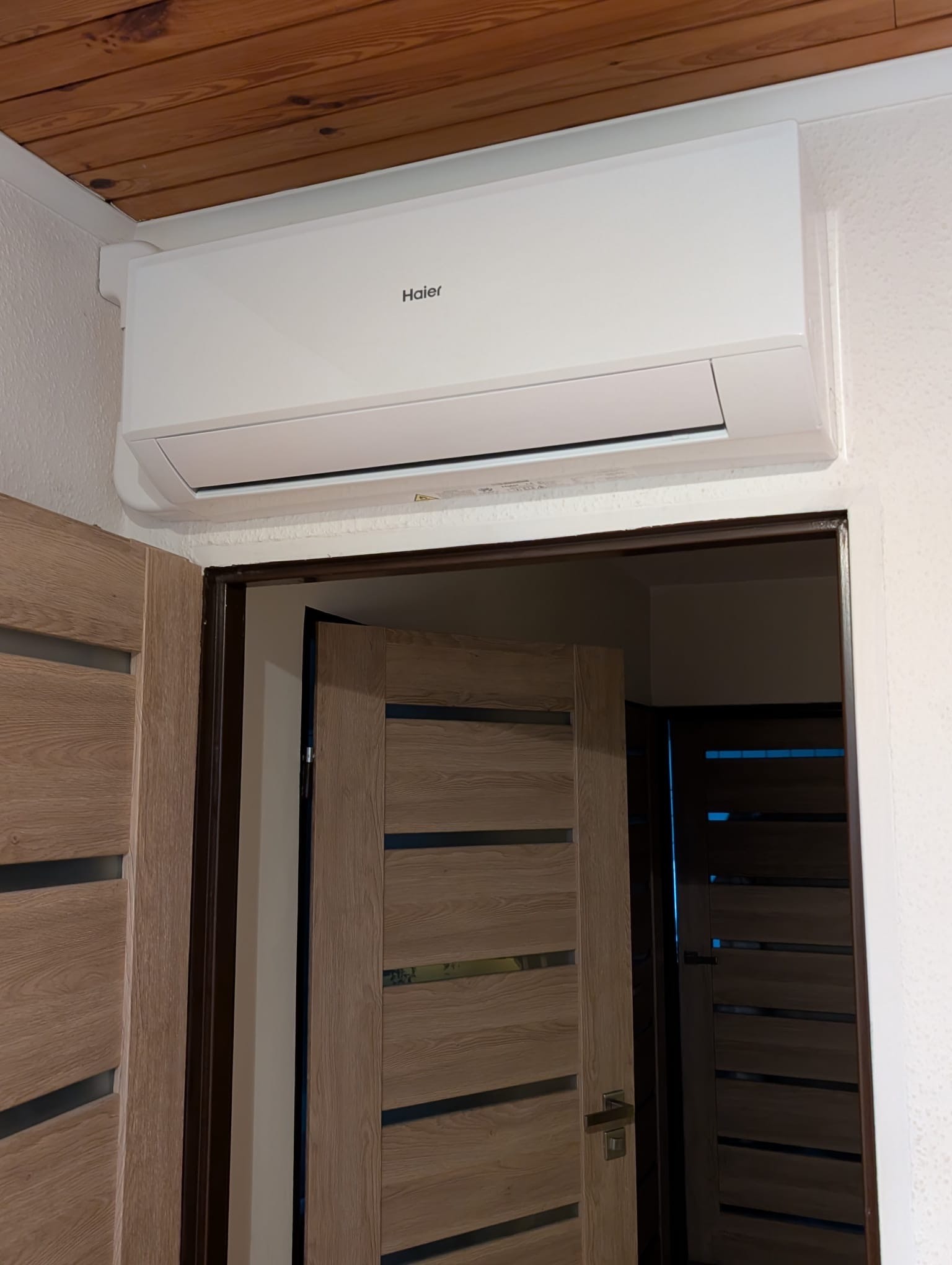 Biała klimatyzacja Haier zamontowana nad drzwiami z drewnianą okleiną na tle ściany o nieregularnej fakturze i drewnianego sufitu.