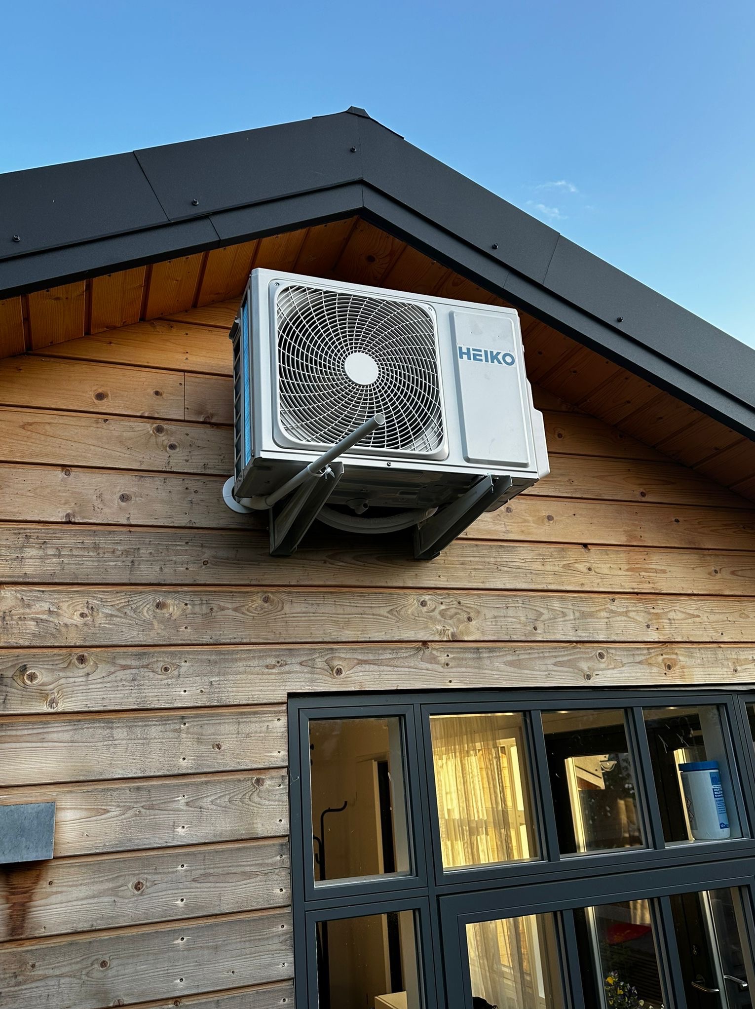 Klimatyzator HEIKO zamontowany na drewnianej elewacji domu, widoczny z boku, na tle błękitnego nieba. Szare wsporniki, czarna rama okna.