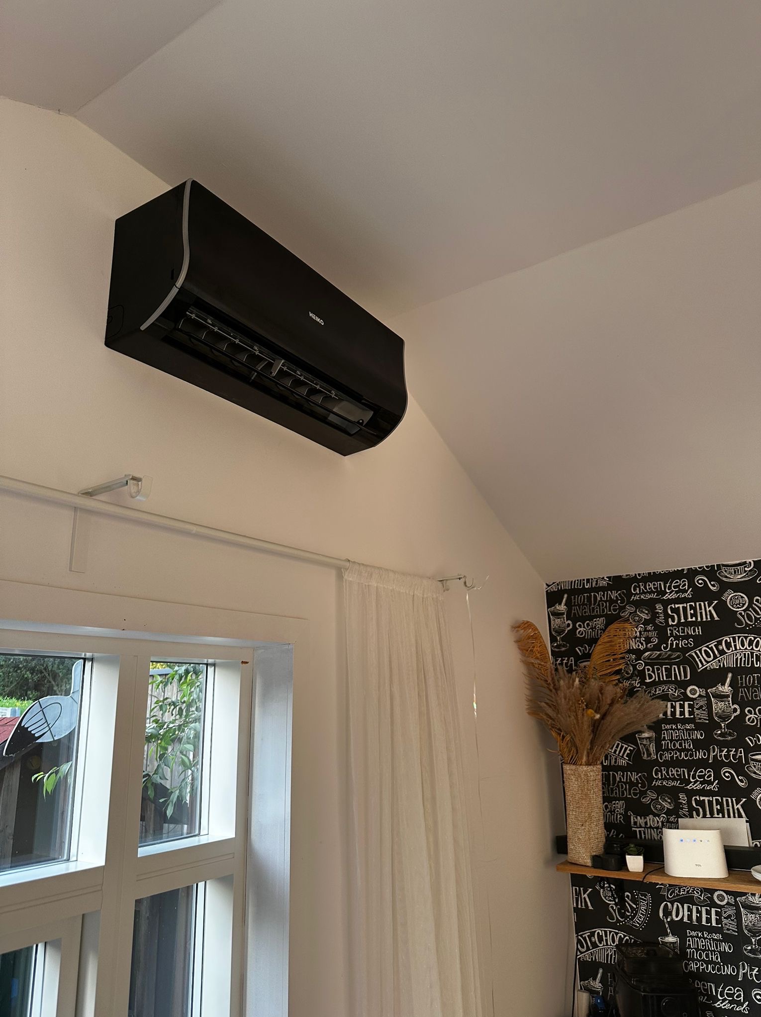 Czarny klimatyzator HEIKO zamontowany na ścianie w pomieszczeniu z oknem i dekoracyjną tapetą. Widoczne detale montażu i estetyczne wykończenie.