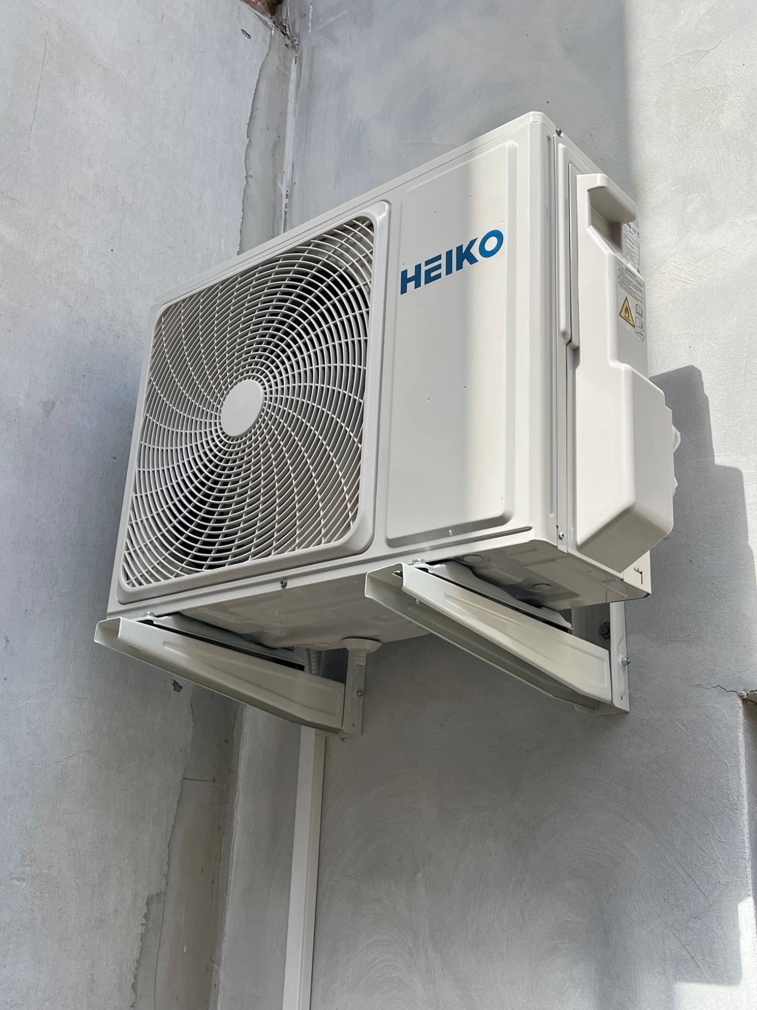 Biała jednostka zewnętrzna klimatyzatora Heiko, zamontowana na szarej, surowej ścianie budynku. Widoczne elementy montażowe i cień rzucany przez urządzenie.