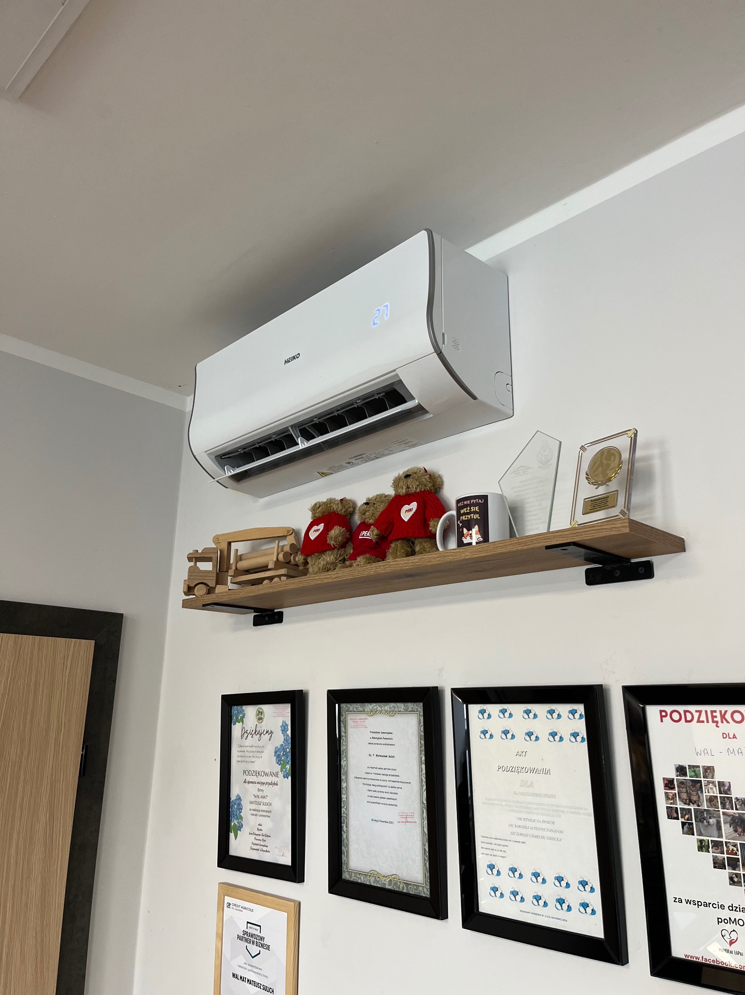 Biała klimatyzacja HEIKO na ścianie nad półką z dekoracjami, w tle ramki z podziękowaniami. Widok z dołu, oświetlenie dzienne, wnętrze pomieszczenia.