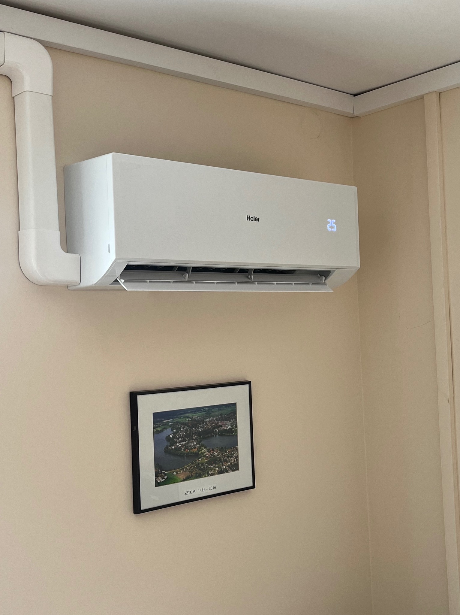 Biały klimatyzator Haier z wyświetlaczem 25°C, zamontowany na beżowej ścianie nad oprawionym zdjęciem Sztumu. Widoczne rury instalacyjne.