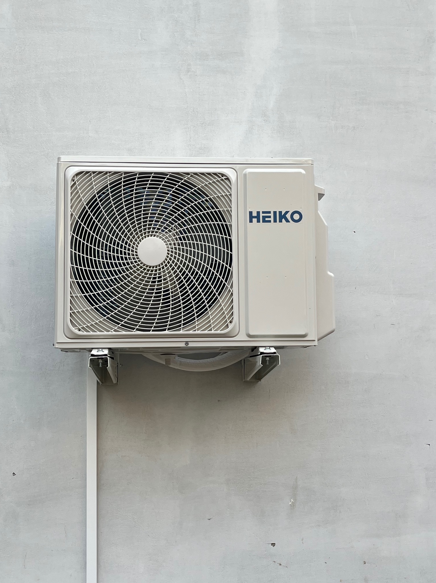 Zamontowana jednostka zewnętrzna klimatyzacji marki HEIKO na szarej, betonowej ścianie. Widoczne metalowe wsporniki i biała listwa maskująca przewody.