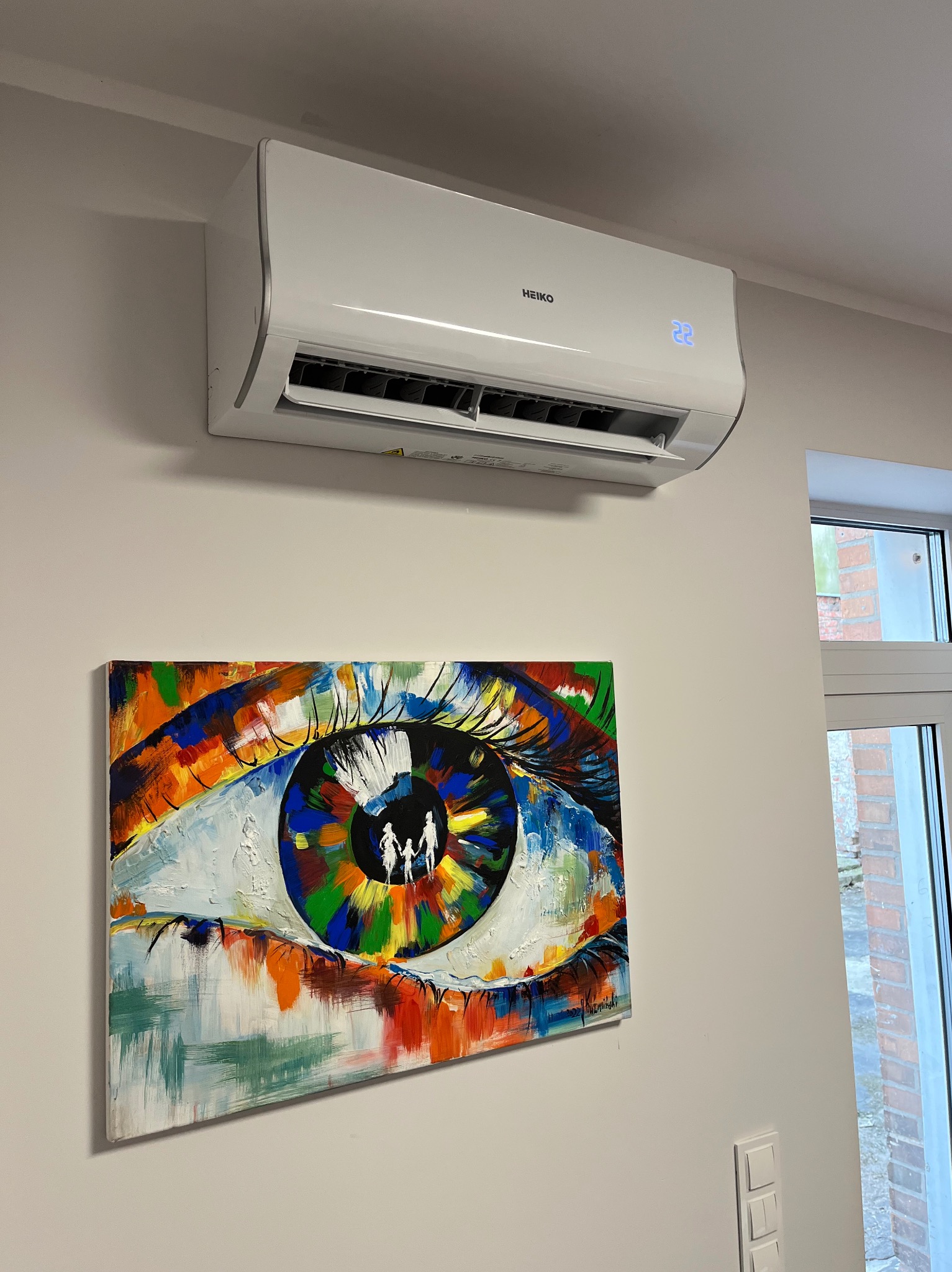 Biała klimatyzacja Heiko na ścianie nad kolorowym obrazem oka. Widoczne okno z ceglanym budynkiem na zewnątrz. Nowoczesny design wnętrza.