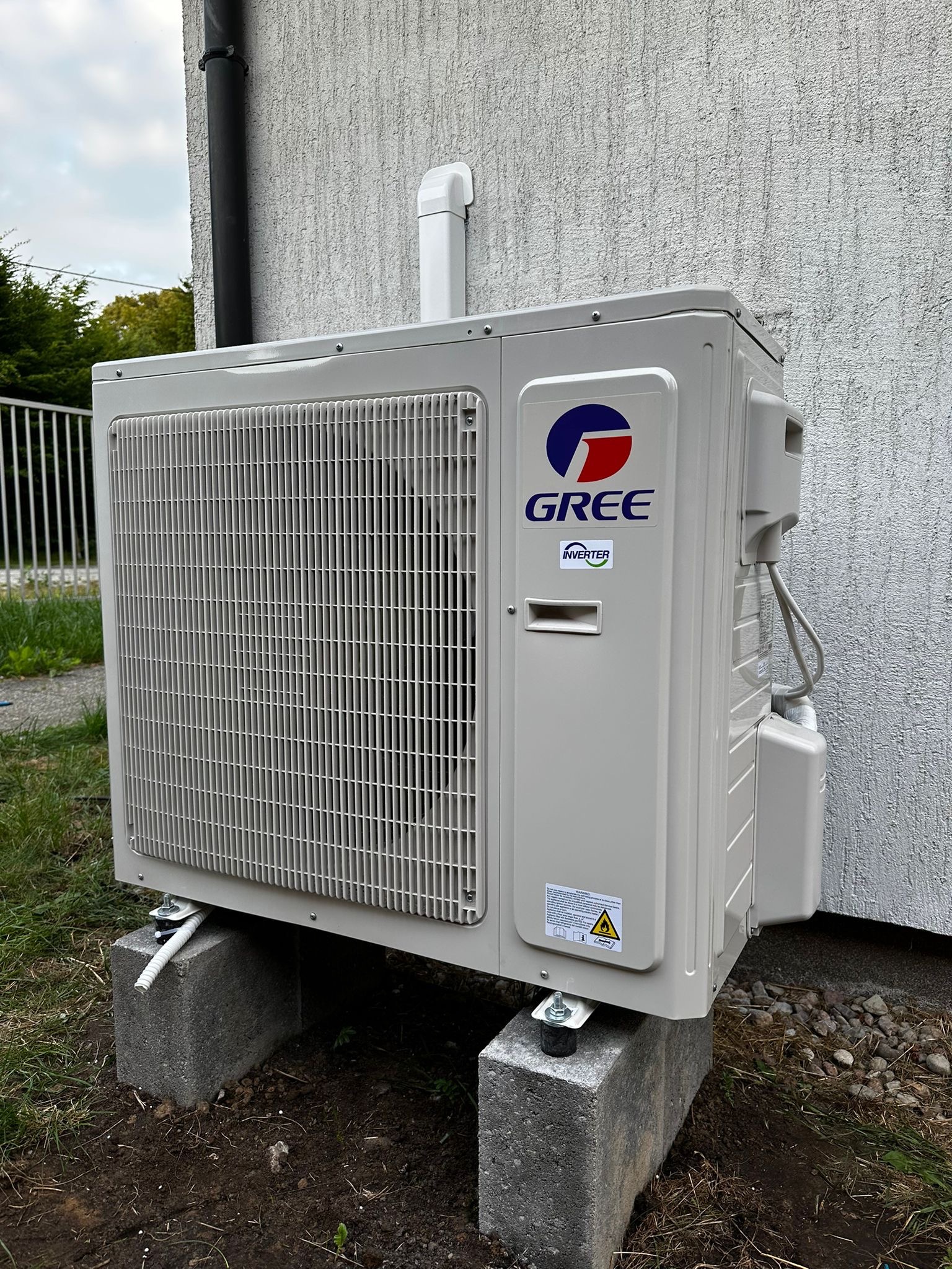 Zamontowana jednostka zewnętrzna klimatyzacji GREE na betonowych bloczkach przy ścianie budynku o chropowatej fakturze. Widoczne elementy instalacji i trawa.
