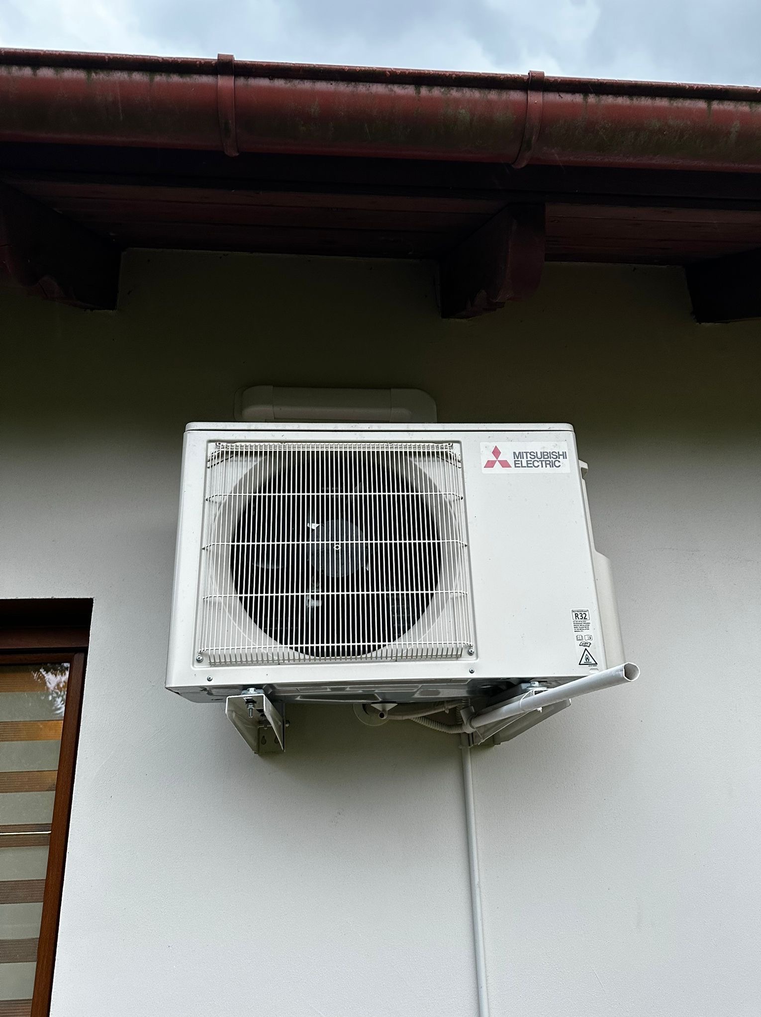 Biała jednostka zewnętrzna klimatyzacji Mitsubishi Electric zamontowana na elewacji budynku z widocznymi elementami instalacji i rynną w tle.
