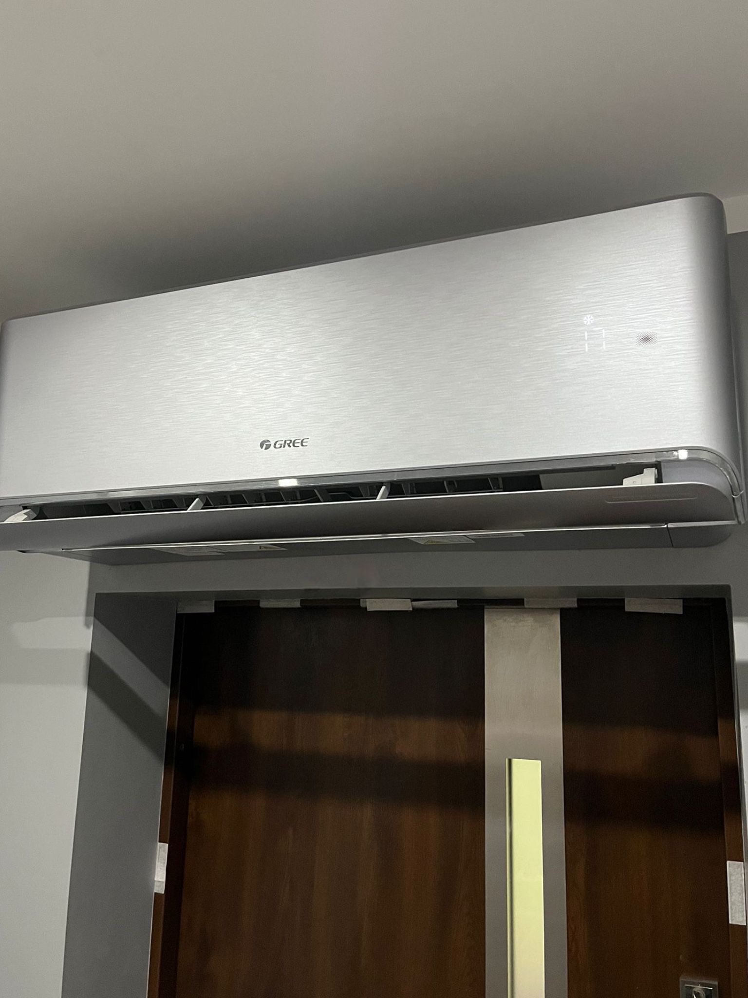 Srebrna klimatyzacja Gree zamontowana nad ciemnymi drzwiami z metalową listwą. Widoczny wyświetlacz temperatury i logo producenta. Minimalistyczny design.