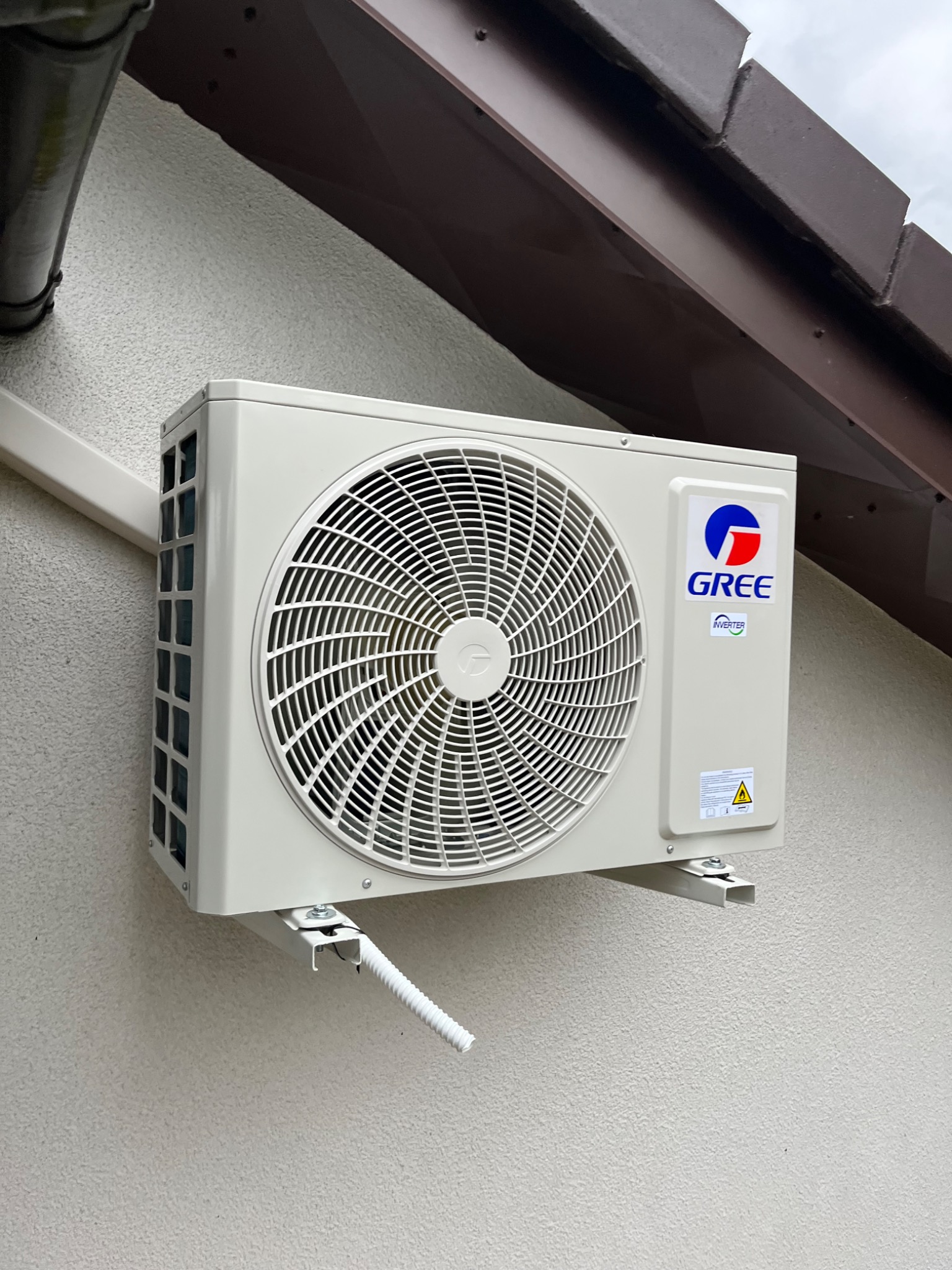 Biała klimatyzacja GREE zamontowana na elewacji budynku z widoczną instalacją odprowadzającą skropliny. Ujęcie z dołu, widoczny fragment dachu.