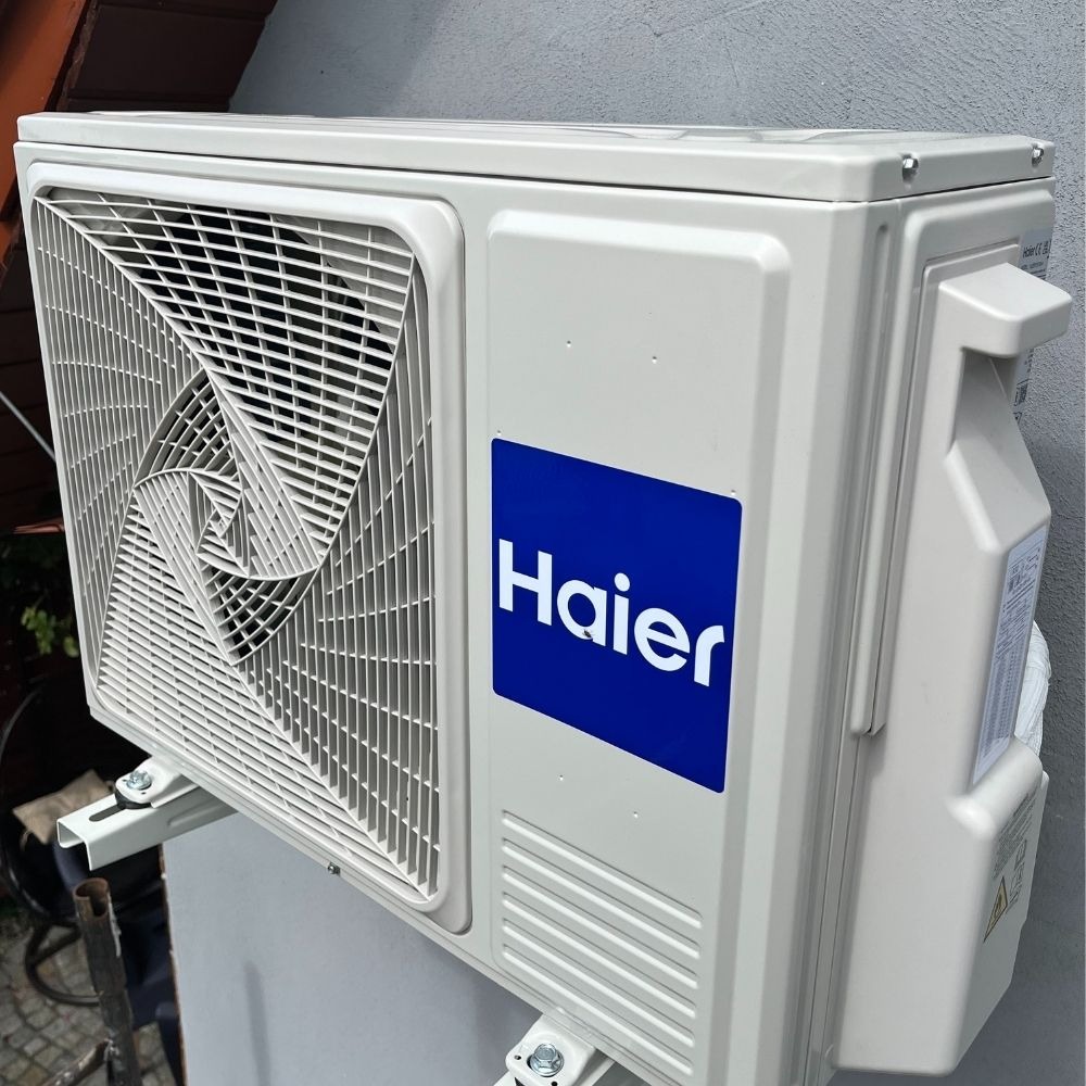 Klimatyzator Haier zamontowany na ścianie, widoczny z boku. Biała obudowa, niebieskie logo i ażurowa kratka wentylacyjna. Widoczne elementy montażowe.