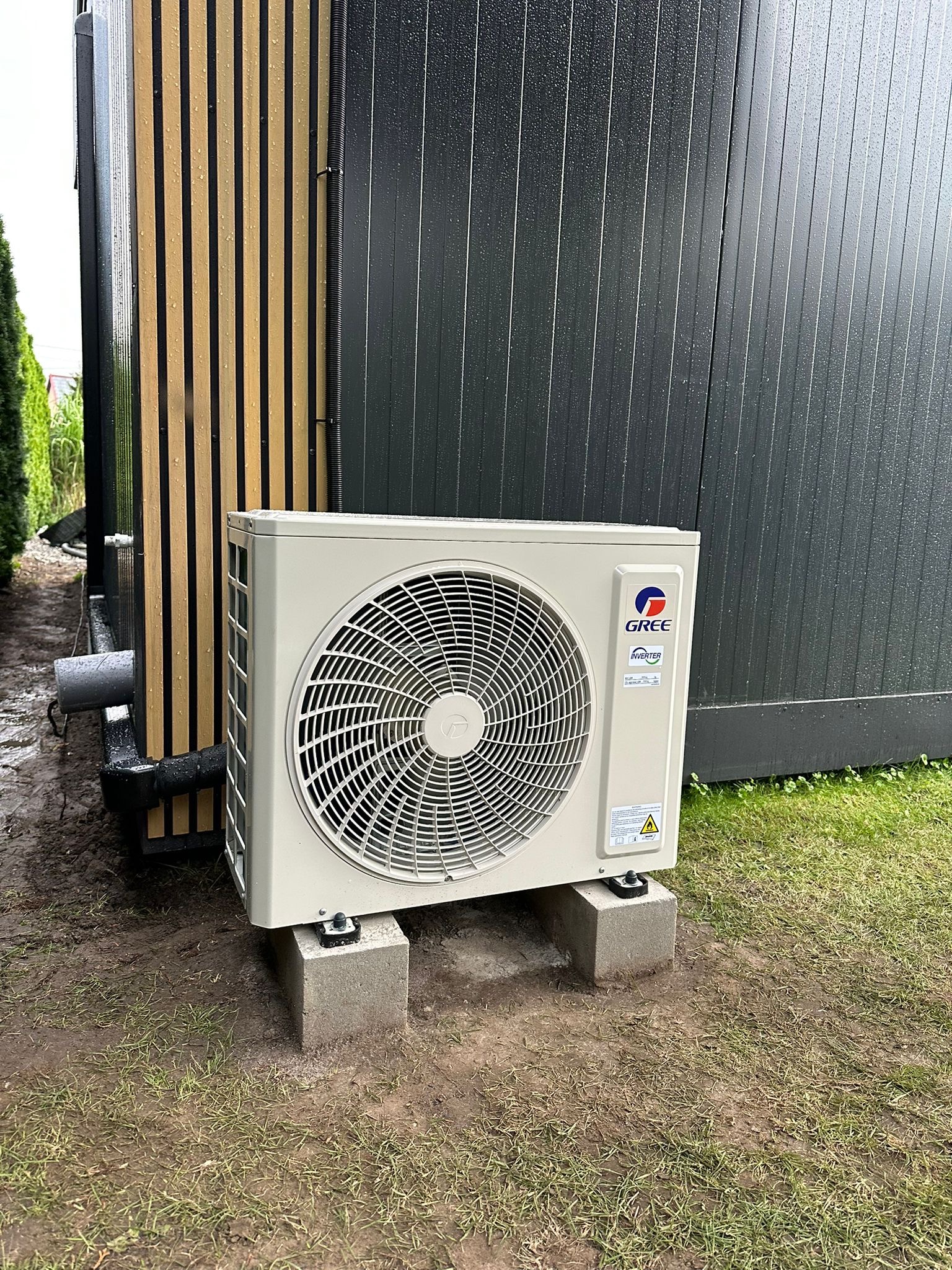 Zewnętrzna jednostka klimatyzacji Gree, umieszczona na betonowych bloczkach obok budynku o nowoczesnej elewacji w deszczowy dzień. Widoczne detale montażu i otoczenie.