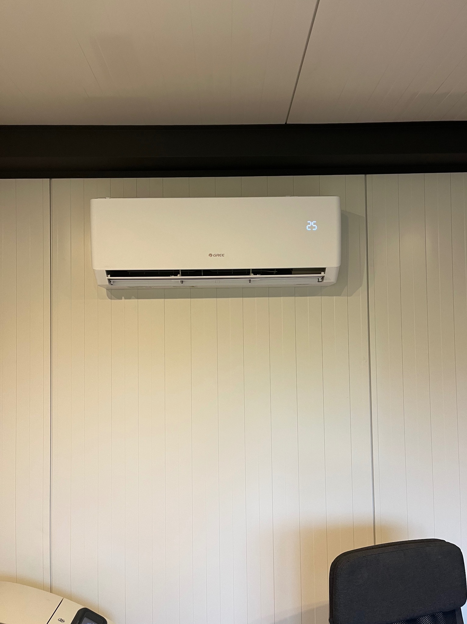 Biały klimatyzator Gree zamontowany na panelowej ścianie, wyświetlający temperaturę 25 stopni Celsjusza, widoczny fragment czarnej belki sufitowej i krzesła biurowego.
