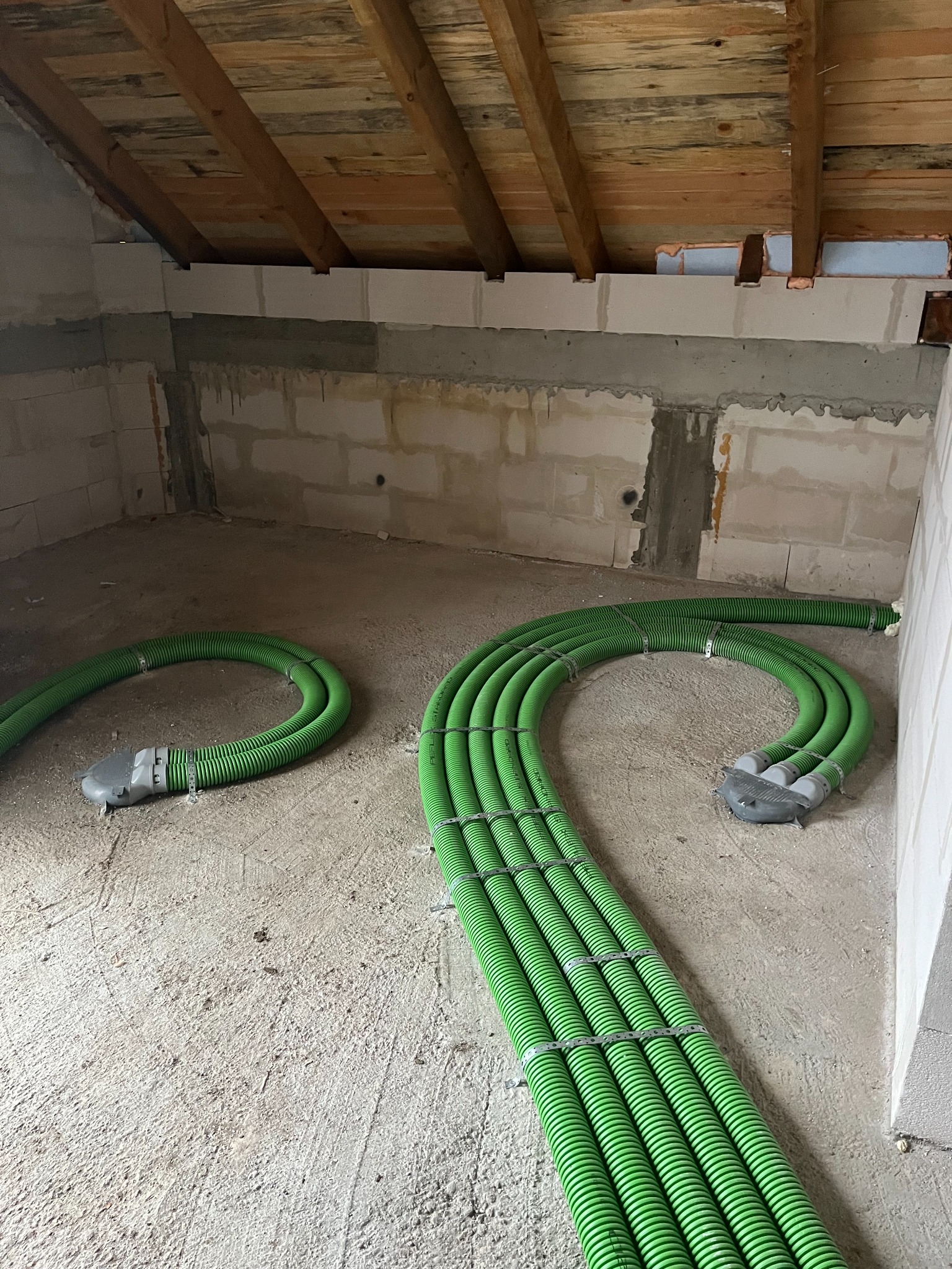 Zielone rury wentylacyjne ułożone na surowym podłożu w pomieszczeniu z surowymi ścianami z bloków i drewnianym stropem. Instalacja w trakcie realizacji.