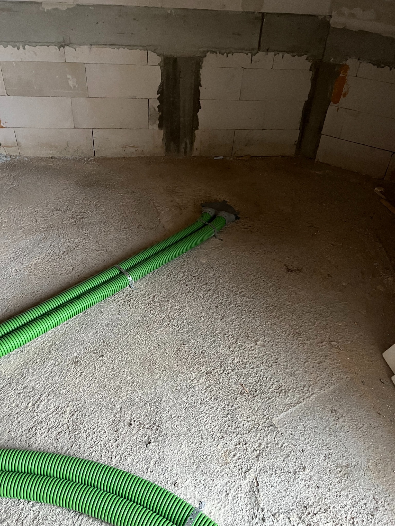 Zielone, karbowane rury wentylacyjne ułożone na surowym betonie wewnątrz budynku w trakcie budowy. Widoczne bloczki betonowe ścian.