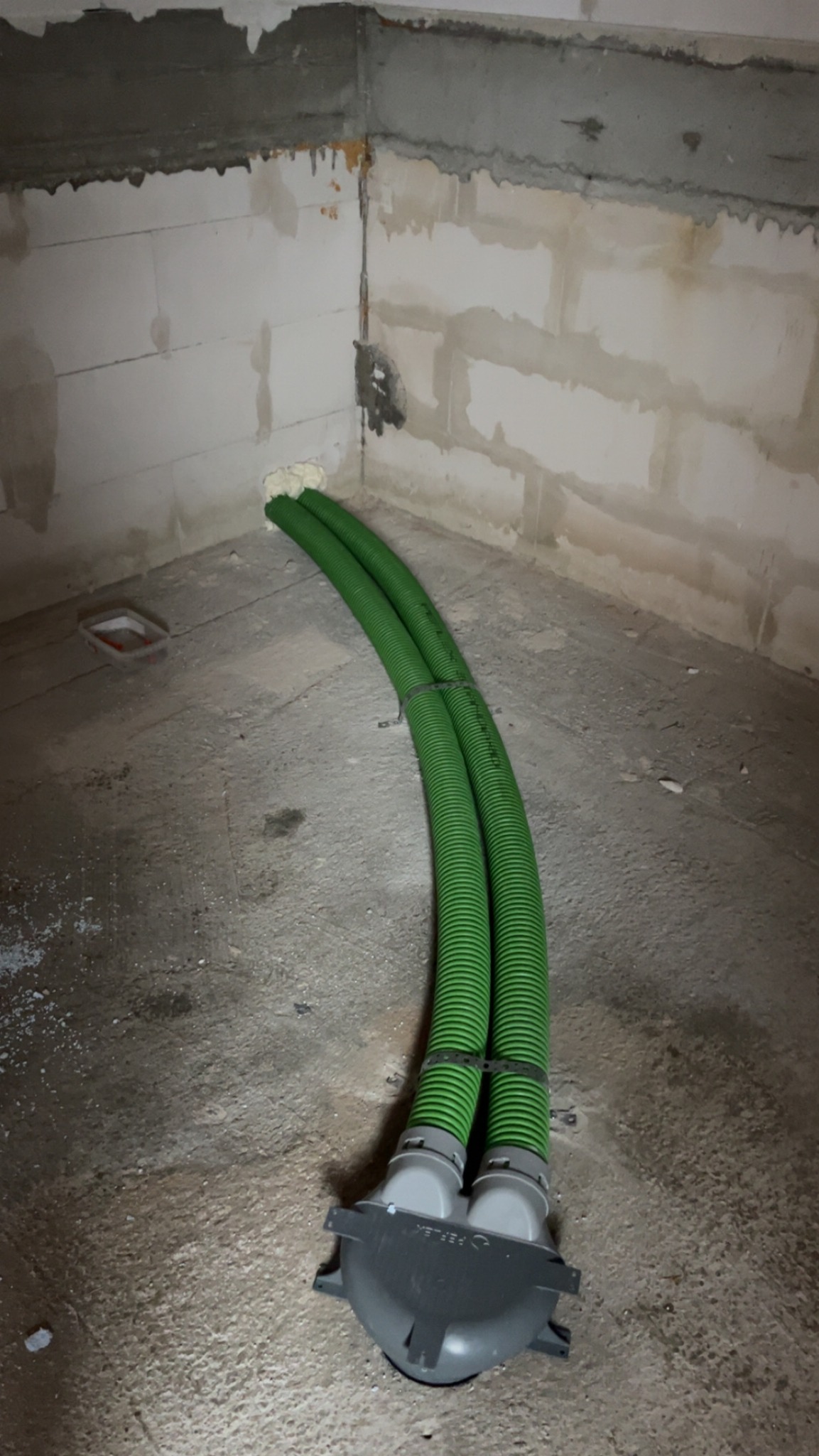 Dwie zielone, karbowane rury wentylacyjne przymocowane do szarego rozdzielacza na surowej podłodze w pomieszczeniu z bloków betonowych. Pianka montażowa uszczelnia przejście rur przez ścianę.