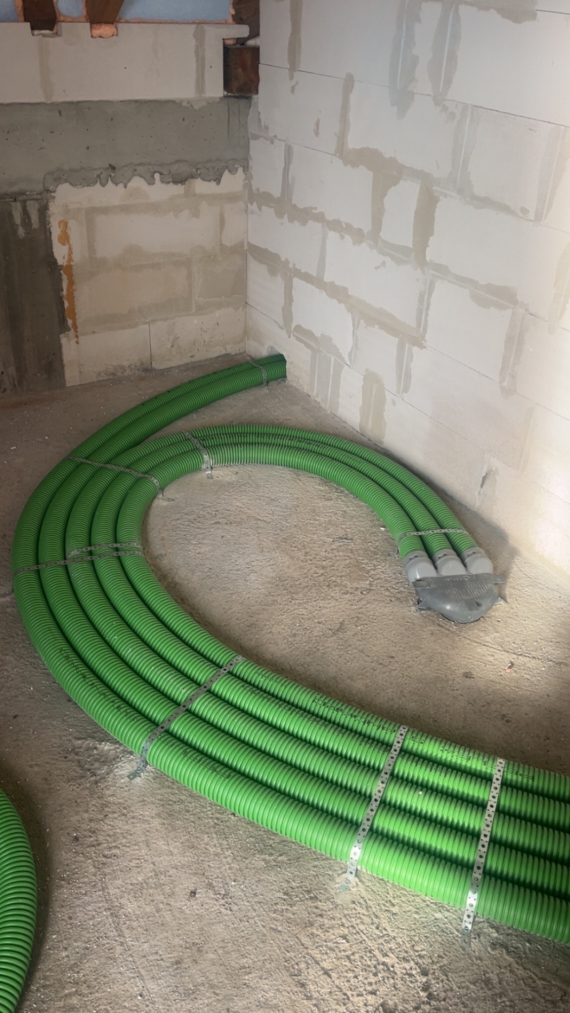 Zielone, karbowane rury wentylacyjne ułożone na podłodze w surowym wnętrzu, przymocowane metalowymi obejmami, prowadzące do otworu w ścianie z białych bloczków.