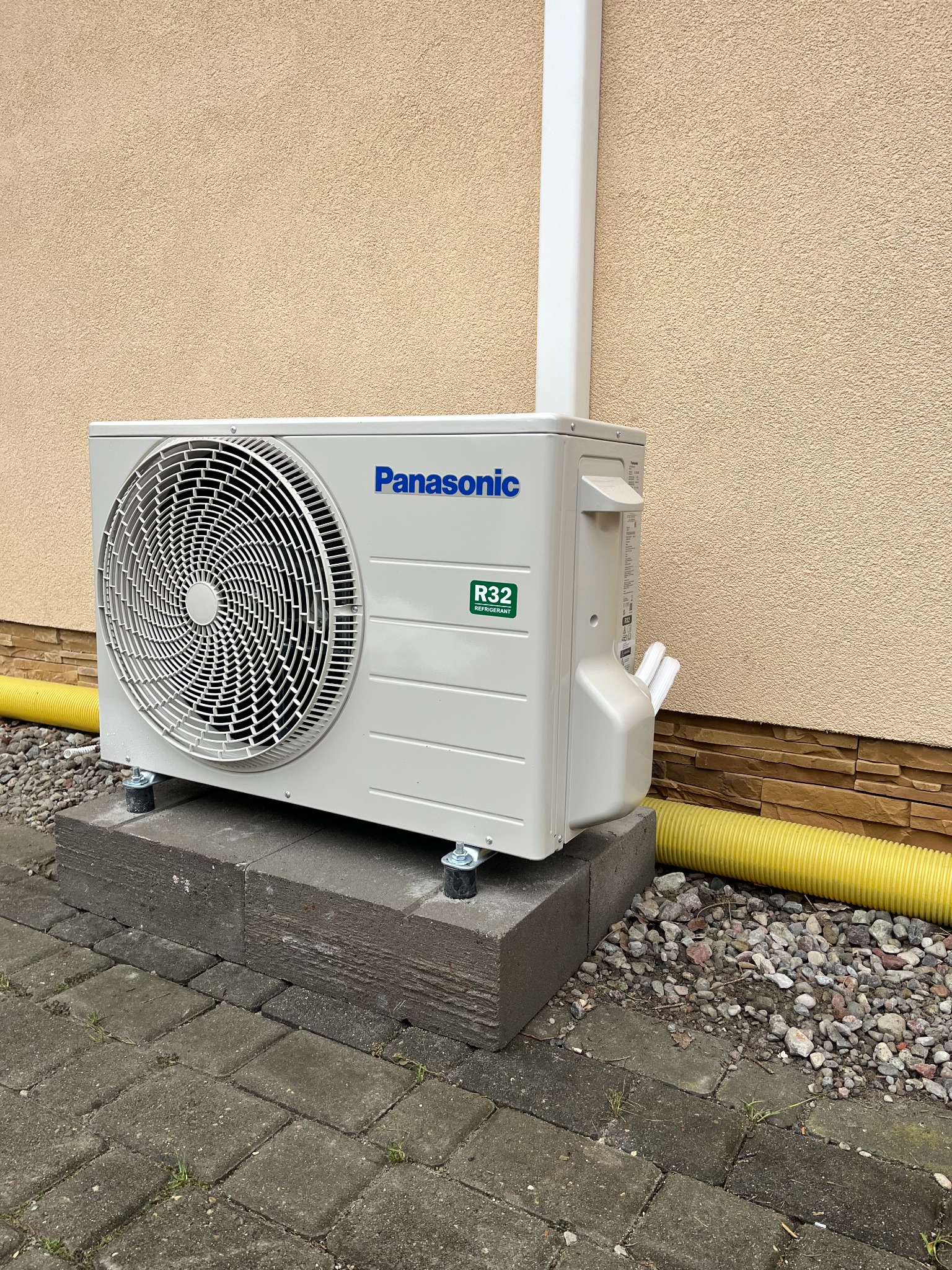 Zewnętrzna jednostka klimatyzacji Panasonic na betonowym fundamencie, z widocznymi rurami odprowadzającymi skropliny i żółtą rurą osłonową, na tle elewacji budynku.